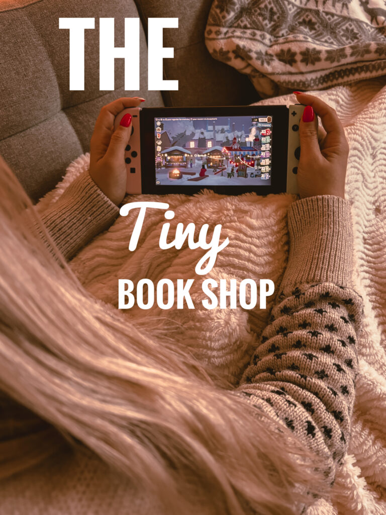Sorbet-Kiwi allongée sur un canapé tient une console Nintendo Switch blanche affichant une scène de marché de Noël sur The Tiny Bookshop. Elle est enveloppée dans une couverture beige et porte un pull à motifs foncés. Une grande mèche de cheveux tombe sur le côté.
