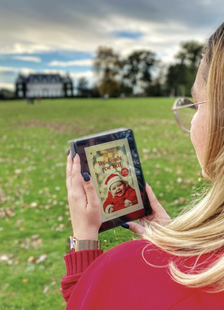 Sorbet-Kiwi de dos lit le roman numérique « Une famille pour Noël » sur une liseuse, dans un parc verdoyant avec un grand château en arrière-plan, par une journée d’automne.