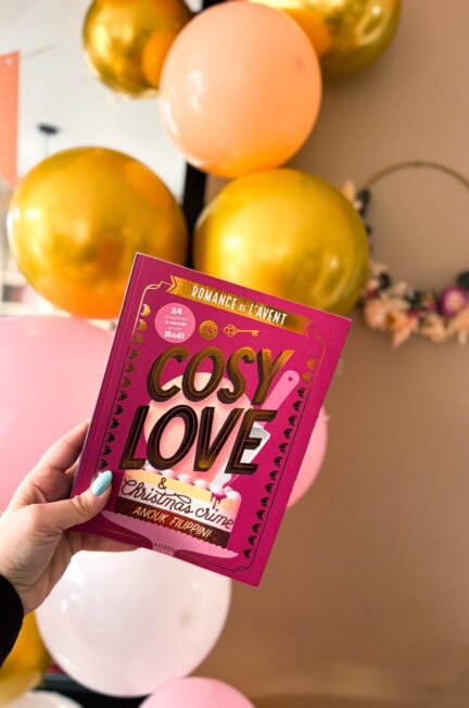Livre Cosy Love & Christmas Crime d’Anouk Filippini tenu à la main devant des ballons roses et dorés, évoquant une ambiance festive et de Noël.
