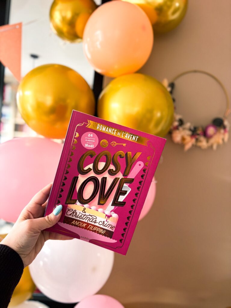 Livre Cosy Love & Christmas Crime d’Anouk Filippini tenu à la main devant des ballons roses et dorés, évoquant une ambiance festive et de Noël.