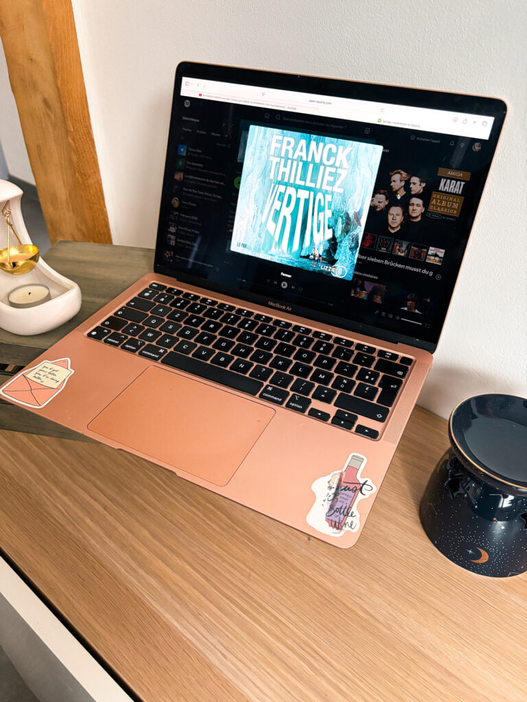 Ordinateur portable rose posé sur un bureau en bois, avec le livre audio Vertige de Franck Thilliez affiché à l’écran sur Spotify. À côté, une bougie allumée et une tasse sombre créent une ambiance cosy, propice à l’écoute et à la concentration.