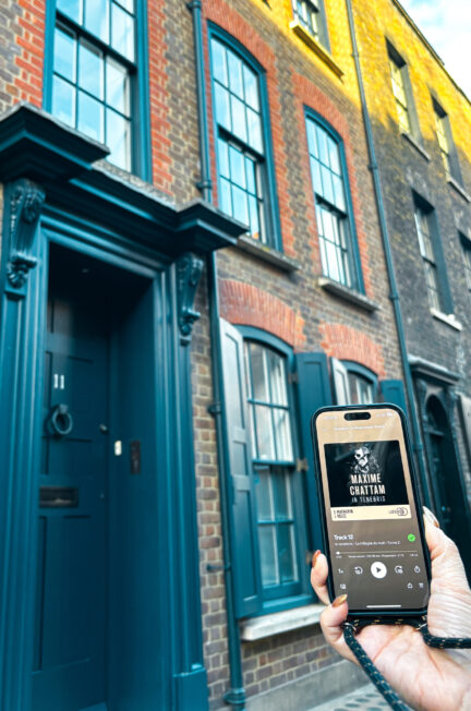 Téléphone affichant l’audiobook In Tenebris de Maxime Chattam devant une façade de maisons en briques aux portes bleues de Whitechapel à Londres, ambiance hivernale.