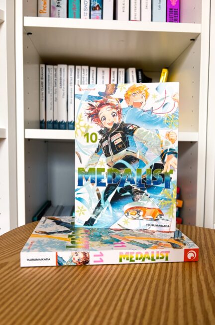 Le tome 10 du manga Medalist Junior-GP de Tsurumaikada est posé sur une table en bois, avec sa couverture illustrant Inori et Tsukasa en pleine dynamique de patinage artistique, devant une bibliothèque blanche.