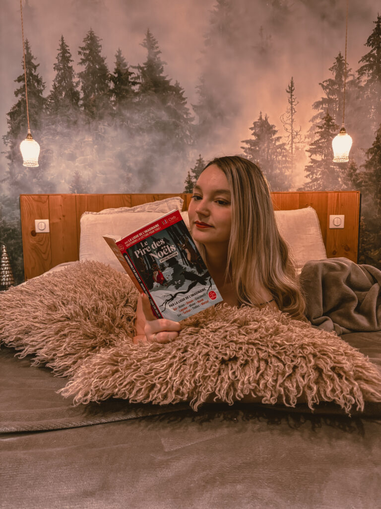 Sorbet-Kiwi est allongée dans un lit douillet, entourée de coussins et d’une décoration hivernale, en train de lire Le pire des Noëls : spécial frayeur dans une ambiance cosy.
