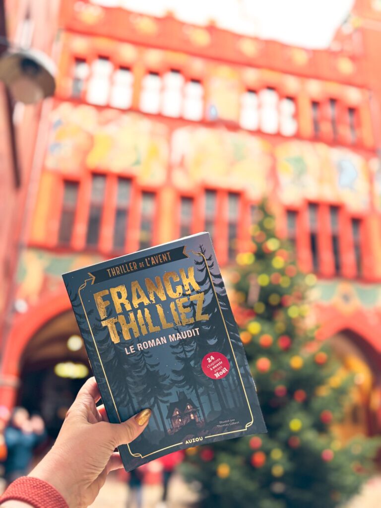 Le roman maudit de Franck Thilliez tenu à la main devant le sapin de Noël de l'hôtel de ville de Bâle, illustrant un thriller jeunesse au format calendrier de l’avent.