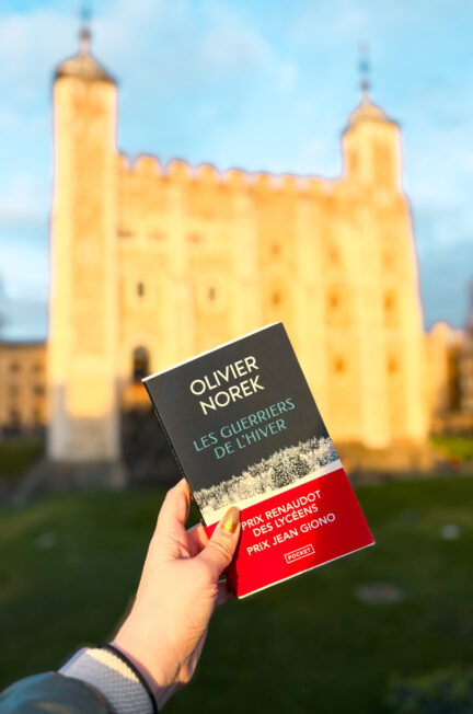 Livre Les guerriers de l’hiver d’Olivier Norek tenu à la main devant la Tour de Londres illuminée, en arrière-plan flou, au crépuscule.