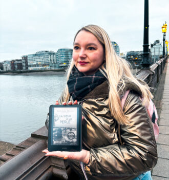 Stéphanie tenant une Kindle affichant La sœur à la perle de Lucinda Riley sur un pont à Battersea, avec la Tamise et des immeubles modernes en arrière-plan à Londres.
