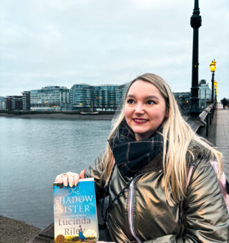 Sorbet-Kiwi tenant The Shadow Sister de Lucinda Riley sur un pont à Battersea, avec la Tamise et les immeubles modernes en arrière-plan à Londres.