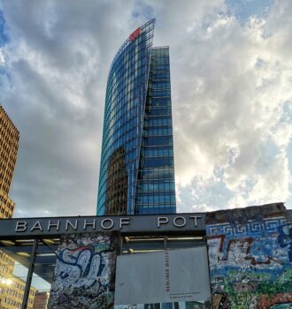 Entrée de la station Bahnhof Potsdamer Platz à Berlin, avec des fragments du mur de Berlin couverts de graffitis au premier plan et une tour moderne en verre de la Deutsche Bahn s’élevant sous un ciel nuageux - ex République démocratique allemande