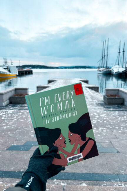 Exemplaire de I’m every woman de Liv Strömquist tenu dans une main gantée devant le fjord d’Oslo, avec le port et des voiliers en arrière-plan sous un ciel hivernal.