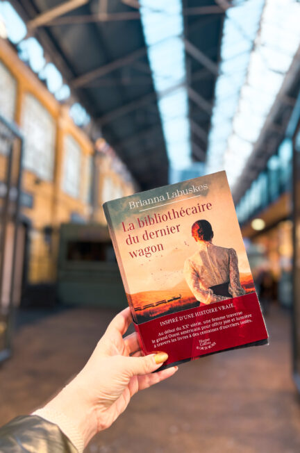 Exemplaire de La bibliothécaire du dernier wagon de Brianna Labuskes tenu en main à Spitalfields Market à Londres, dans une halle lumineuse aux structures métalliques en arrière-plan flou.