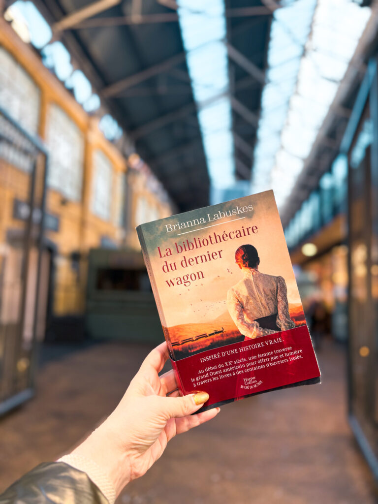 Exemplaire de La bibliothécaire du dernier wagon de Brianna Labuskes tenu en main à Spitalfields Market à Londres, dans une halle lumineuse aux structures métalliques en arrière-plan flou.