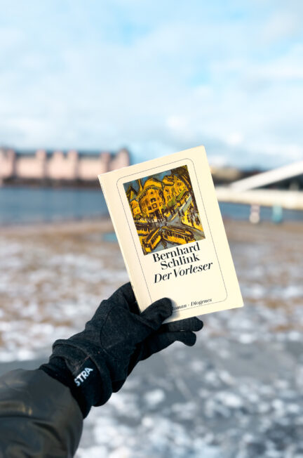 Exemplaire allemand de Der Vorleser, titre allemand de Le liseur, de Bernhard Schlink tenu dans une main gantée devant l’opéra d’Oslo, avec la mer et les bâtiments en arrière-plan hivernal.