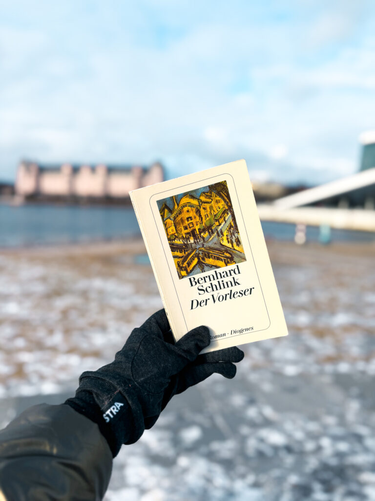Exemplaire allemand de Der Vorleser, titre allemand de Le liseur, de Bernhard Schlink tenu dans une main gantée devant l’opéra d’Oslo, avec la mer et les bâtiments en arrière-plan hivernal.
