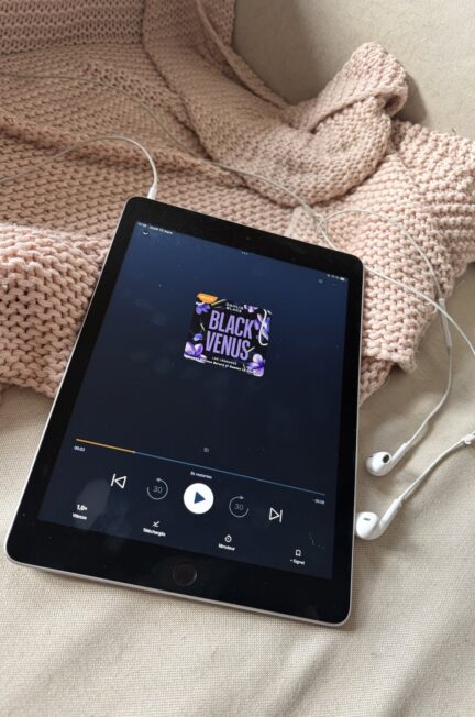 Une tablette affiche la couverture audio du livre Black Venus de Dahlia Blake, posée sur un plaid beige avec des écouteurs blancs à côté. L’écran montre le lecteur en pause, prêt pour une séance d’écoute.