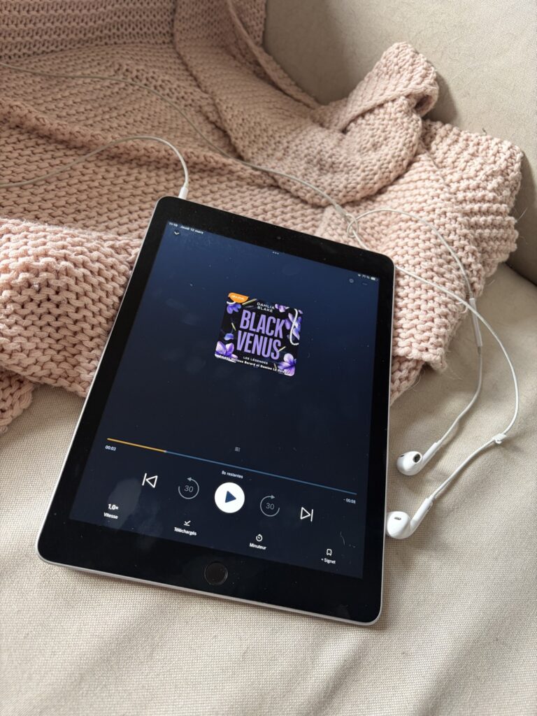 Une tablette affiche la couverture audio du livre Black Venus de Dahlia Blake, posée sur un plaid beige avec des écouteurs blancs à côté. L’écran montre le lecteur en pause, prêt pour une séance d’écoute.
