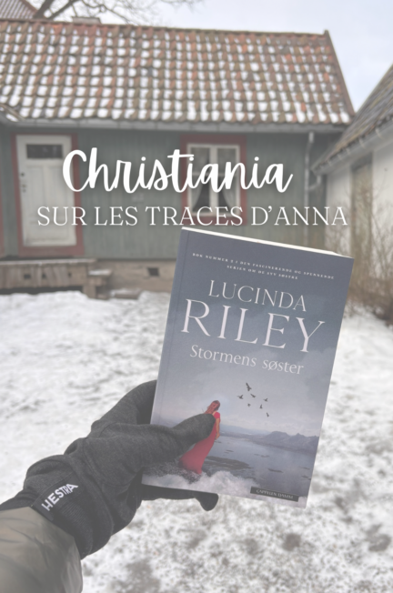 Une main gantée tient le livre Stormens søster de Lucinda Riley devant une maison en bois typique de Christiania, enneigée. Le texte « Christiania Sur les traces d’Anna » est superposé en blanc.