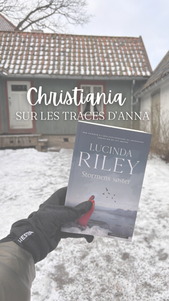 Une main gantée tient le livre Stormens søster de Lucinda Riley devant une maison en bois typique de Christiania, enneigée. Le texte « Christiania Sur les traces d’Anna » est superposé en blanc.