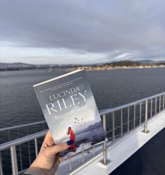 Une main gantée présente le livre Stormens søster de Lucinda Riley depuis un pont métallique d'un bateau enneigé, avec une vue sur le fjord gelé d’Oslo. Une ambiance hivernale immersive, sur les traces d’Anna.