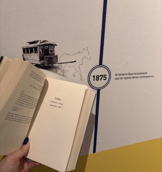 Une main tient ouvert un livre de Lucinda Riley devant une frise historique du musée d’Oslo, datée de 1875 et illustrant un tram tiré par un cheval. Une plongée dans l’époque d’Anna, sur les traces d’Anna.