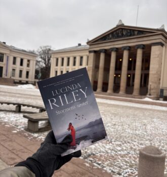 Une main gantée tient le livre Stormens søster devant l’entrée de l'université enneigée, typique de l’architecture de Christiania. Une exploration urbaine et littéraire, sur les traces d’Anna.