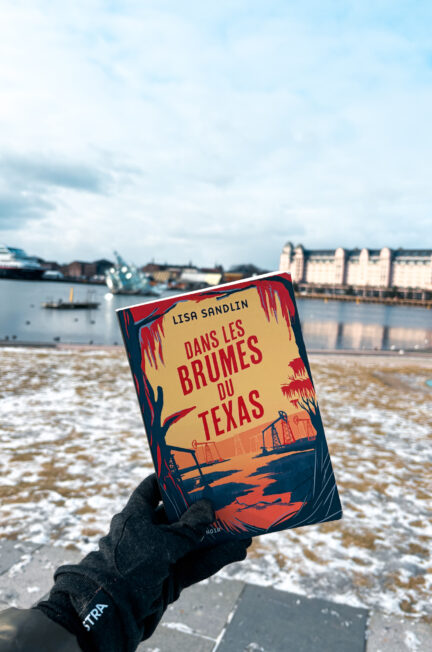 Une main gantée tient le livre Dans les brumes du Texas de Lisa Sandlin, couverture visible, devant le port d'Oslo enneigé avec des bâtiments industriels et des bateaux en arrière-plan.