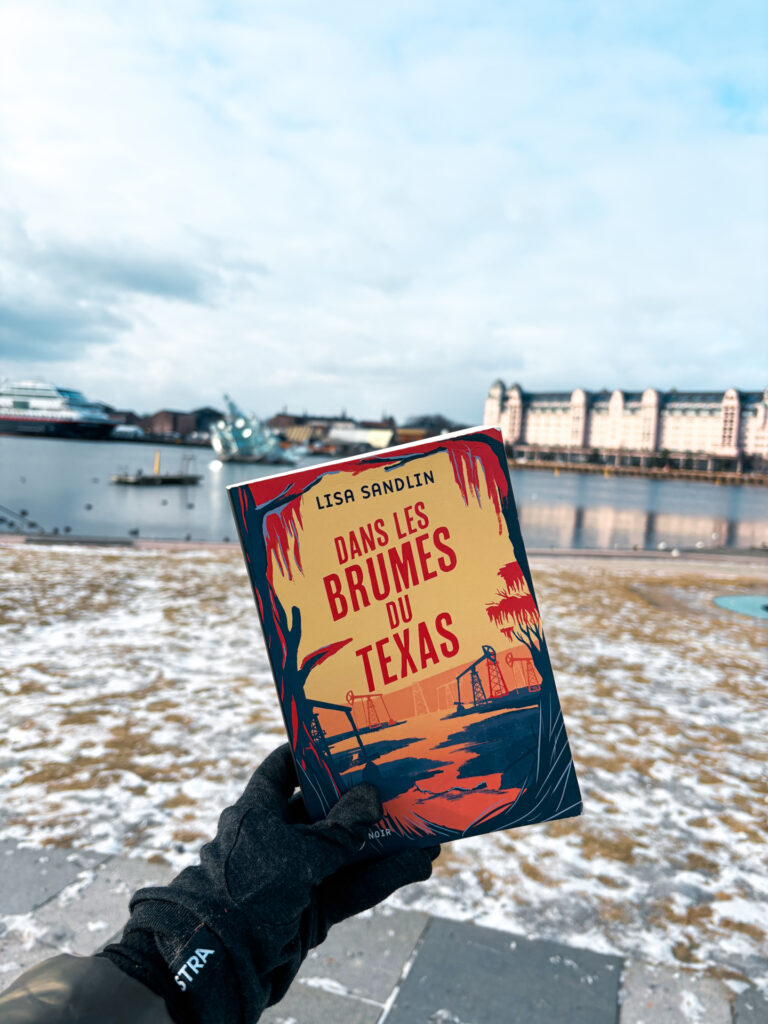 Une main gantée tient le livre Dans les brumes du Texas de Lisa Sandlin, couverture visible, devant le port d'Oslo enneigé avec des bâtiments industriels et des bateaux en arrière-plan.