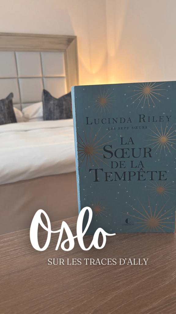 Le livre La Sœur de la tempête de Lucinda Riley posé sur une table en bois, avec en arrière-plan un lit et une tête de lit capitonnée du Grand Hotel d'Oslo. Le texte « Oslo Sur les traces d’Ally » est superposé en blanc.