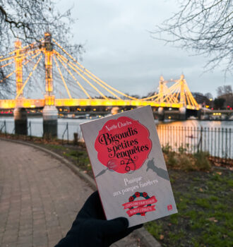 Une main tient le livre Panique aux pompes funèbres de Naëlle Charles, couverture visible, devant le pont illuminé d’Albert Bridge à Londres, en arrière-plan.