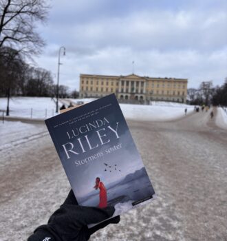 Une main gantée tient le livre Stormens søster de Lucinda Riley, couverture visible, devant un paysage enneigé et un bâtiment jaune clair, le Palais royal, à Oslo. Un moment de lecture immersif, sur les traces d’Ally.