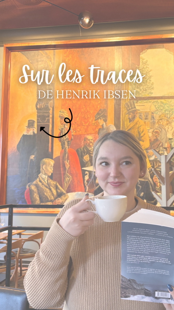 Sorbet-Kiwi sourit en tenant une tasse de café et le livre Stormens søster de Lucinda Riley, devant la fresque représentant Henrik Ibsen au Grand Café. Le texte « Sur les traces de Henrik Ibsen » est superposé en blanc.