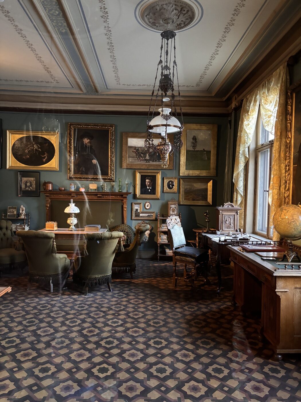 Intérieur du bureau d’Henrik Ibsen au musée Ibsen à Oslo, avec des murs décorés de motifs géométriques, des étagères remplies de livres, de portraits encadrés et d’objets d’époque. Une lampe suspendue ornée de perles et un globe terrestre sont visibles.