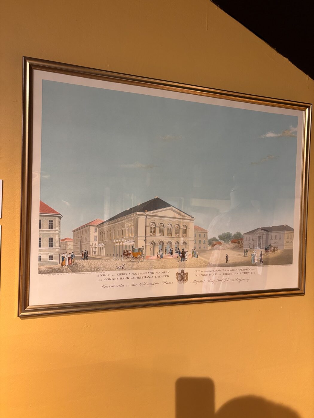 Une gravure encadrée représentant le théâtre de Christiania et la banque de Norvège au XIXe siècle, exposée sur un mur jaune clair du musée d'Oslo. La gravure montre des bâtiments classiques et des personnages en costume d’époque.