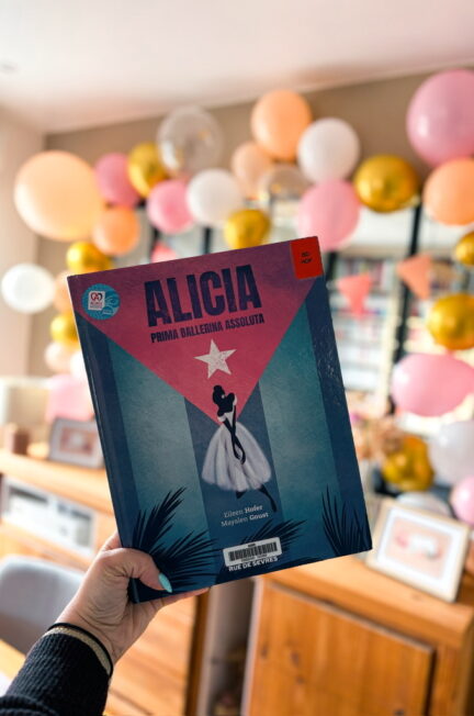 livre Alicia Prima Ballerina Assoluta tenu à la main devant une verrière avec décoration de ballons colorés en arrière plan