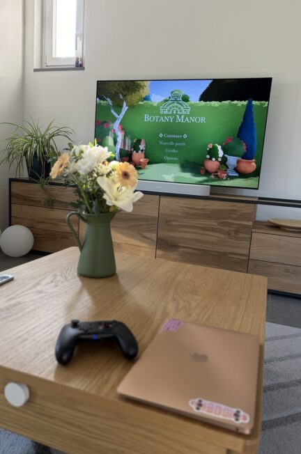 Un MacBook, une manette de switch et un bouquet de fleurs printanières dans un vase vert sont posés sur une table basse en bois devant une TV affichant l'écran d'accueil du jeu Botany Manor.