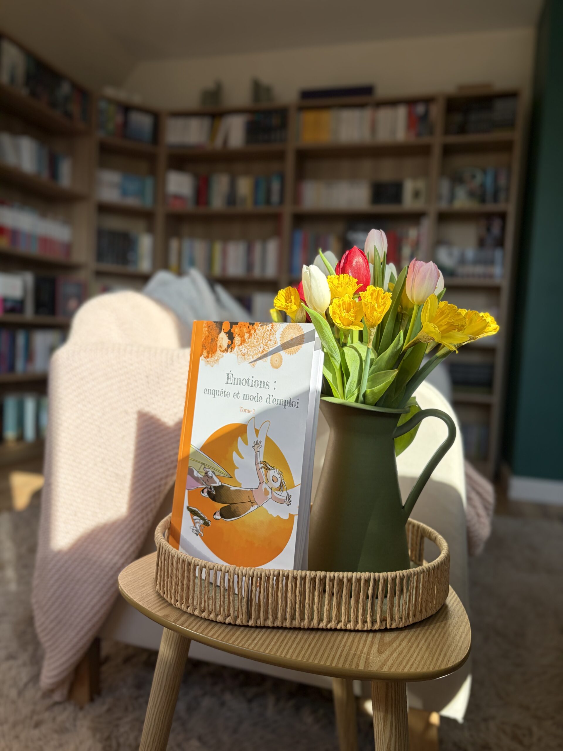 Un livre intitulé Émotions : enquête et mode d'emploi, Tome 1 posé sur une table basse en bois clair, à côté d'un vase vert contenant un bouquet de tulipes jaunes, rouges et blanches. En arrière-plan, un canapé avec une couverture rose et des étagères remplies de livres.