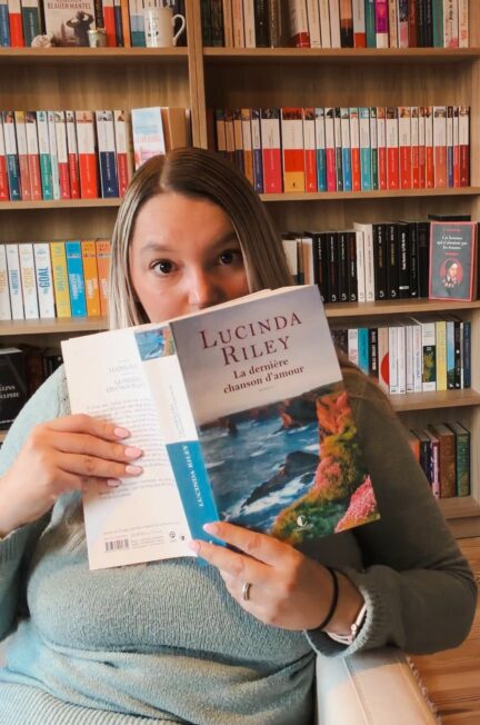 Personne assise dans un fauteuil devant une grande bibliothèque, tenant le roman La dernière chanson d’amour de Lucinda Riley ouvert devant son visage, ne laissant apparaître que ses yeux. Elle porte un pull vert et un jean, et la scène se déroule dans un intérieur chaleureux dédié à la lecture.