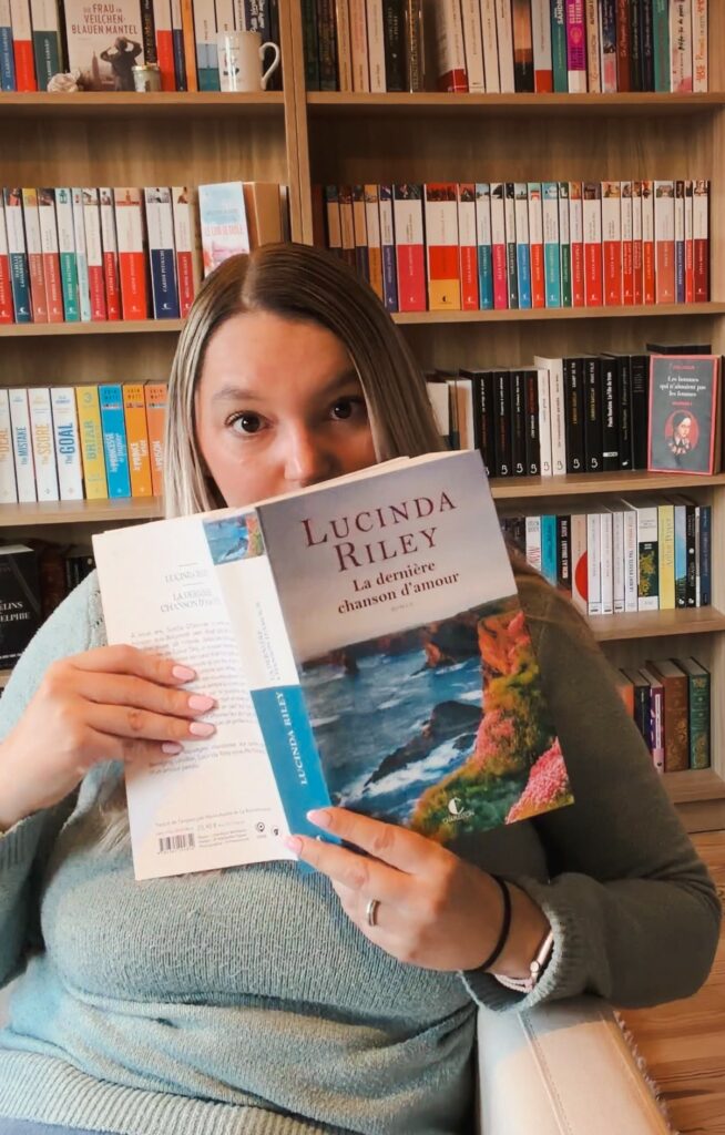 Personne assise dans un fauteuil devant une grande bibliothèque, tenant le roman La dernière chanson d’amour de Lucinda Riley ouvert devant son visage, ne laissant apparaître que ses yeux. Elle porte un pull vert et un jean, et la scène se déroule dans un intérieur chaleureux dédié à la lecture.