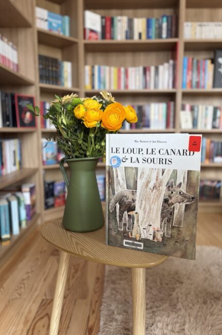 Le loup, le canard et la souris de Mac Barnett et Jon Klassen sur une petite table en bois avec un vase vers et des fleurs du printemps devant une bibliothèque remplie de livres