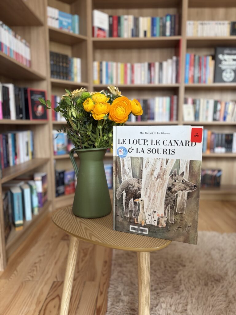 Le loup, le canard et la souris de Mac Barnett et Jon Klassen sur une petite table en bois avec un vase vers et des fleurs du printemps devant une bibliothèque remplie de livres