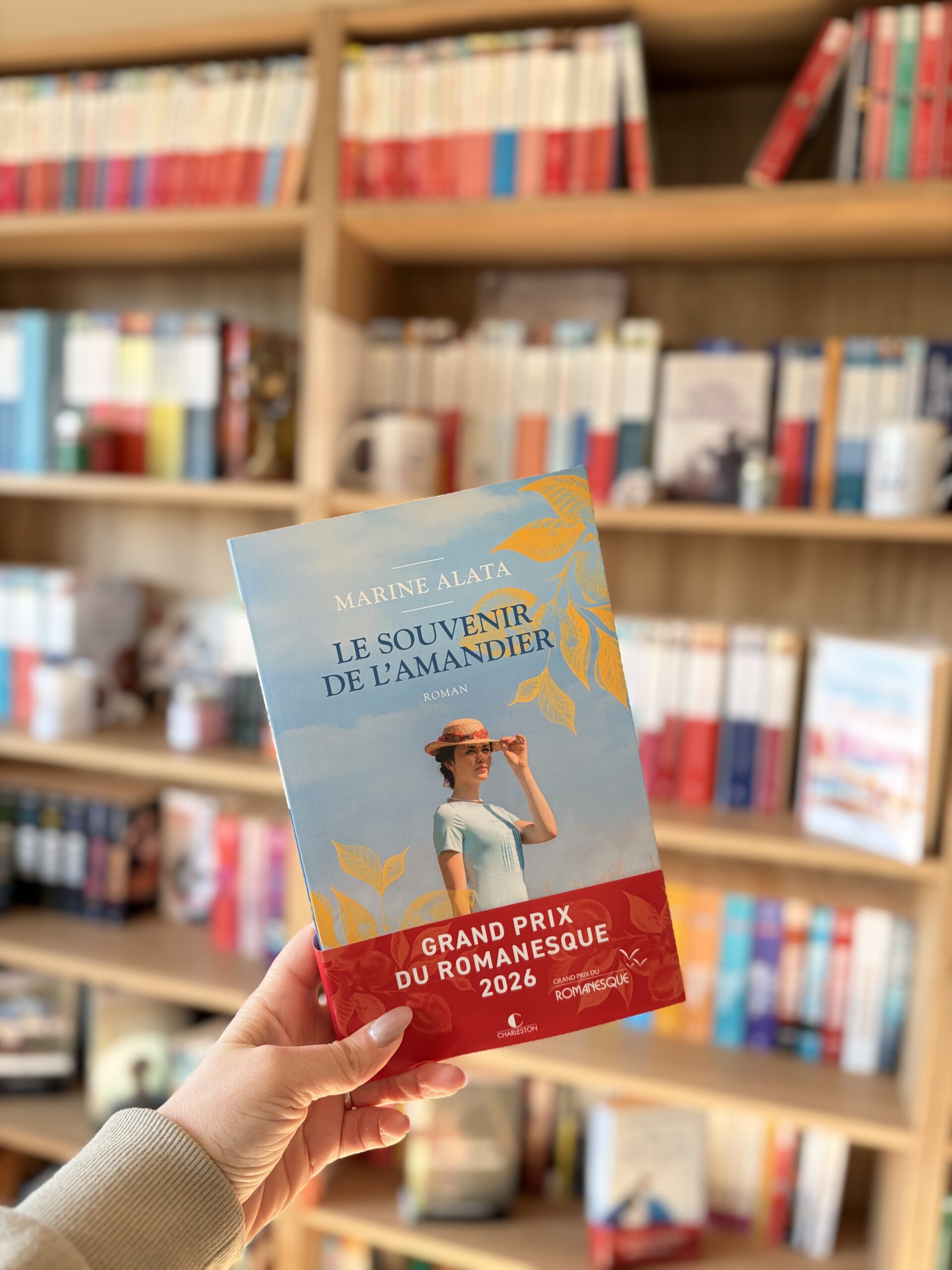 Une main tient le livre Le souvenir de l'amandier de Marine Alata devant une bibliothèque remplie de livres variés. La couverture du livre est visible, avec une illustration représentant une femme en chapeau de paille dans un champ d'amandiers sous un ciel bleu. Le livre porte également la mention "Grand Prix du Romanesque 2026".