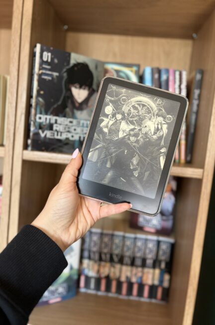 Main tenant une liseuse Kindle affichant l’illustration d’Omniscient Reader’s Viewpoint, devant une bibliothèque en bois remplie de mangas et de romans, dont un volume de la série visible en arrière-plan
