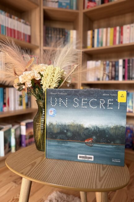 album jeunesse Un secret posé ur une table en bois avec un vase de fleurs séchées devant une bibliothéue remplie de livres