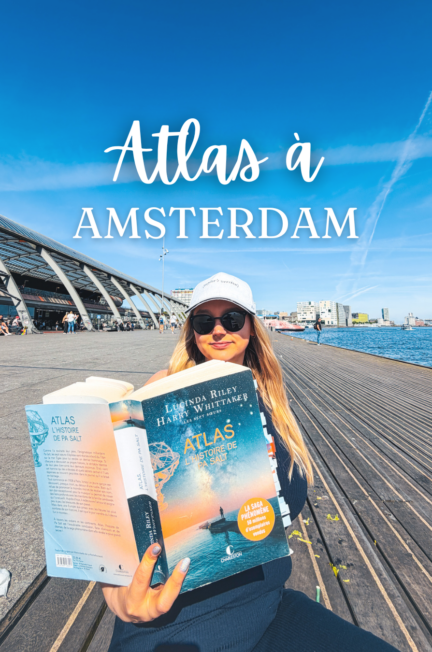 Une lectrice tient le roman Atlas, l’histoire de Pa Salt devant Amsterdam Centraal sous un ciel bleu ensoleillé à Amsterdam.