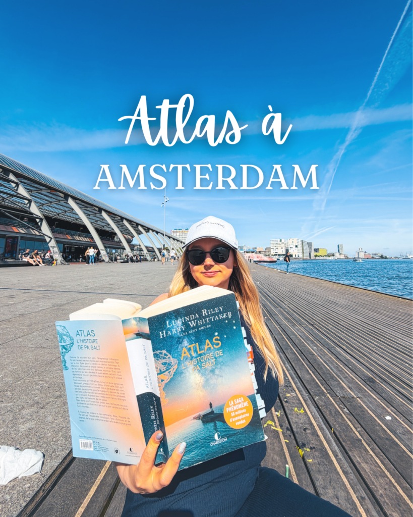 Une lectrice tient le roman Atlas, l’histoire de Pa Salt devant Amsterdam Centraal sous un ciel bleu ensoleillé à Amsterdam.