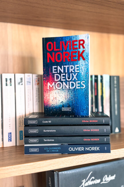 Le roman Entre deux mondes d’Olivier Norek posé sur une pile de livres de l’auteur devant une bibliothèque en bois.