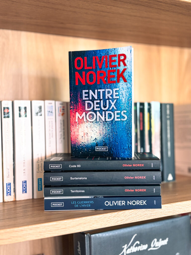Le roman Entre deux mondes d’Olivier Norek posé sur une pile de livres de l’auteur devant une bibliothèque en bois.