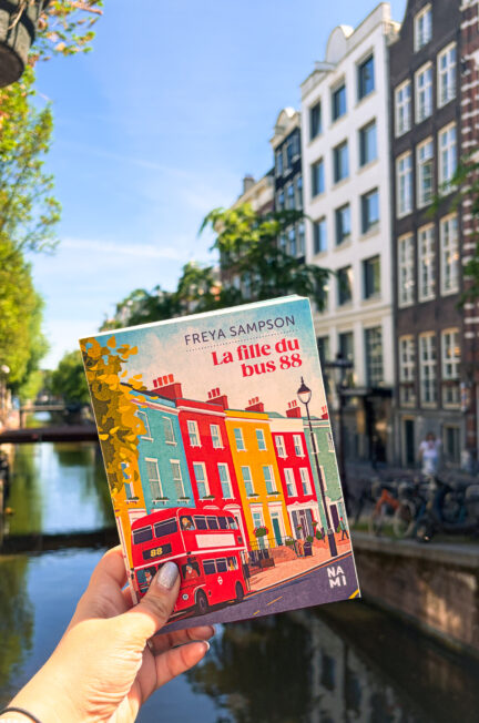 Le roman La fille du bus 88 de Freya Sampson tenu devant un canal ensoleillé d’Amsterdam, avec les façades typiques de la ville en arrière-plan.