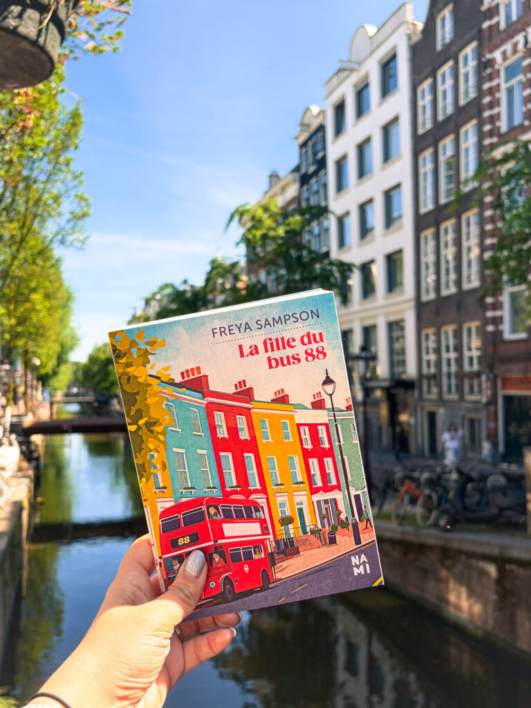 Le roman La fille du bus 88 de Freya Sampson tenu devant un canal ensoleillé d’Amsterdam, avec les façades typiques de la ville en arrière-plan.