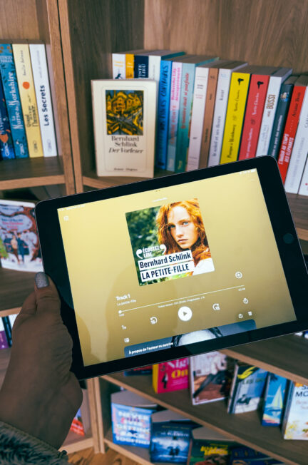 Tablette affichant le livre audio La petite-fille de Bernhard Schlink devant une bibliothèque en bois remplie de romans, avec l’édition allemande Der Vorleser visible en arrière-plan.